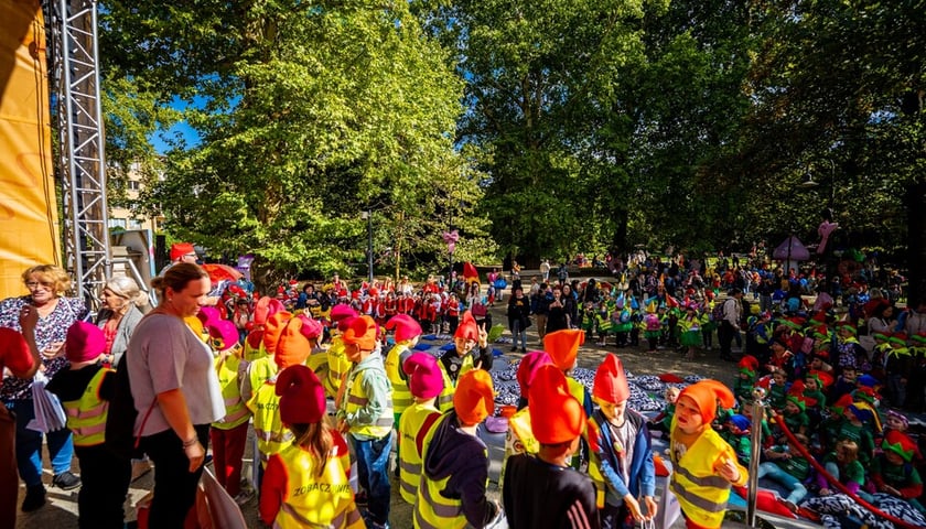 Liczna grupa dzieci w odblaskowych kamizelkach i pomarańczowych czapkach krasnali gromadzi się w parku przy plenerowej scenie podczas Festiwalu Krasnali