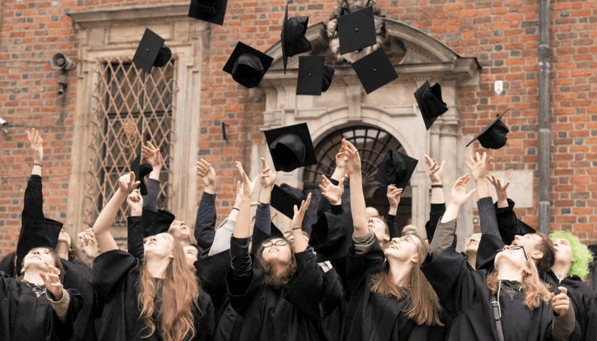 Grupa uśmiechniętych studentów w czarnych togach jednocześnie podrzuca w górę swoje birety studenckie