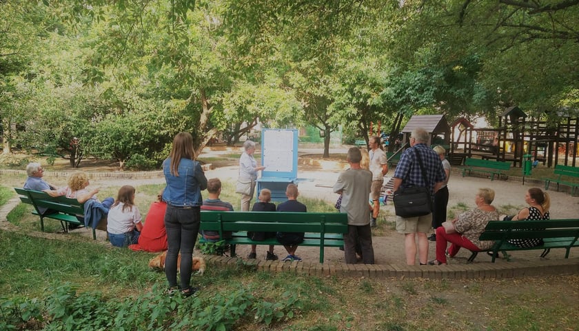 Skwerek otoczony murkiem. Po środku placyku jest piaskownica i drewniana budowla do zabawy. Na środku zgromadzeni mieszkańcy uczestniczą w konsultacjach i prezentacji koncepcji zagospodarowania placu zabaw.