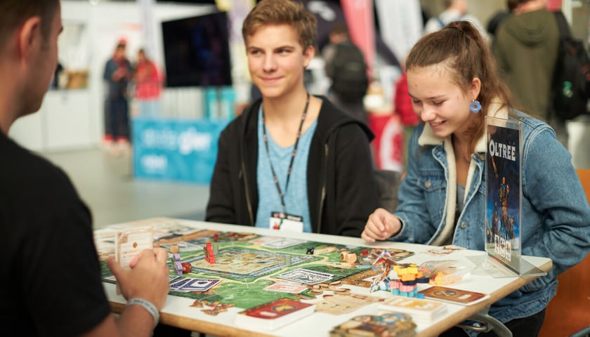 Wrocław Games Week. Wiele z gier planszowych pokazywanych w Hali Stulecia, można wypróbować na miejscu. Na zdjęciu widać trójkę graczy siedzących przy stoliku z grą planszową.