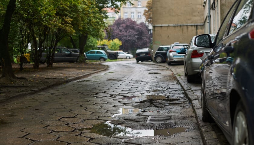 Zaniedbana, dziurawa, podwórkowa ulicę z betonowych płyt. Po prawej przy budynku parkujące w samochody. Po lewej skwer i parkujące wśród drzew samochody. 