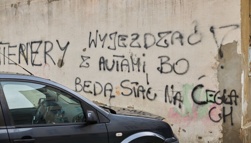 Na zdjęciu bok zaparkowanego pod ścianą samochodu. Na ścianie napis: "wyjeżdżać z autami bo będą stały na cegłach"