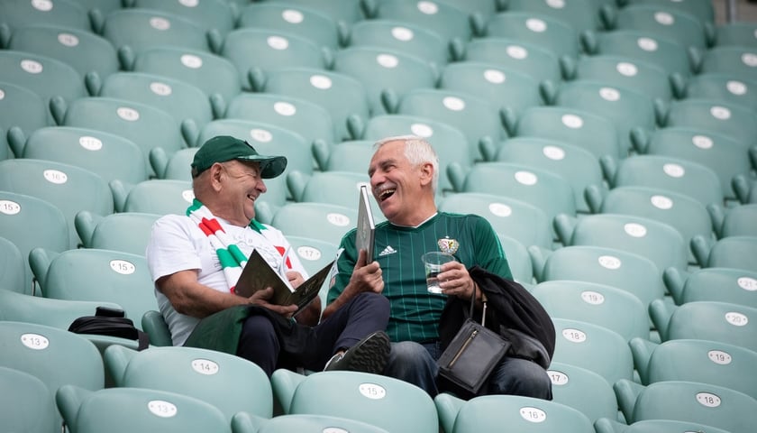 na zdjęciu dwóch kibiców-seniorów na zielonych krzesełkach na trybunie stadionu