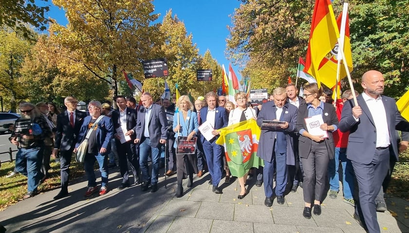 Na zdjęciu grupa samorządowców idąca chodnikiem z flagami 
