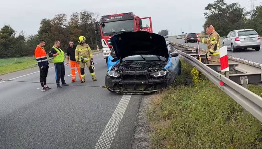 na zdjęciu niebieskie auto BMW M3 G80 z rozbitą maską wyciągane spod barier rozdzielających jezdnię