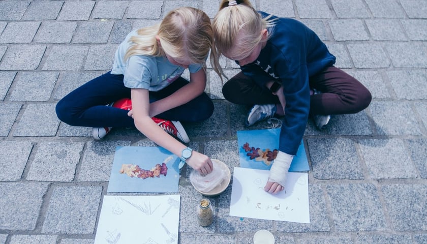 Na zdjęciu dwie dziewczynki siedzące na chodniku, pochylone nad pracami plastycznymi. Zdjęcie ilustracyjne