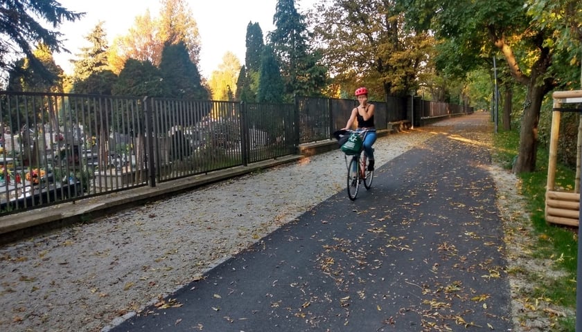 Na zdjęciu istniejący odcinek Promenady Krzyckiej, wzdłuż ogrodzenia cmentarza Ducha Świętego przy ul. Bardzkiej. Do końca roku powstanie jego przedłużenie w kierunku ul. Ziębickiej.  Na trasie kobieta w czarnym podkoszulku jedzie na rowerze.