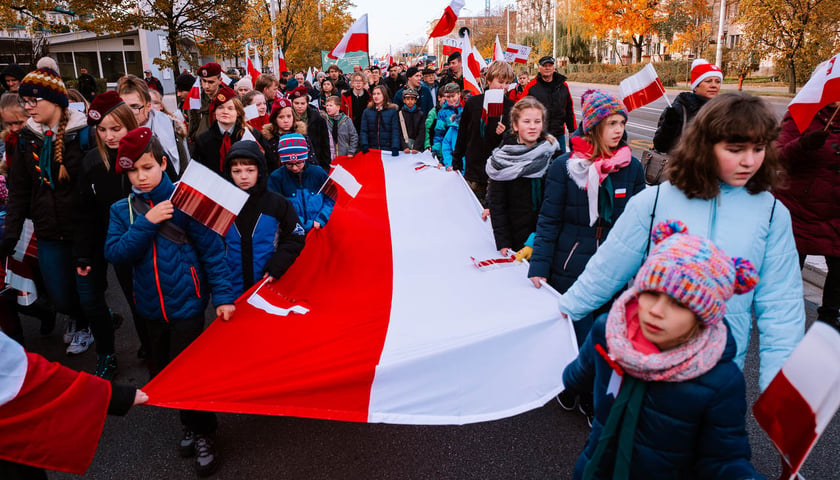 Na zdjęciu uczestnicy Radosnej Parady Niepodległości. Kobiety, mężczyźni i dzieci trzymają dużą biało-czerwoną flagę