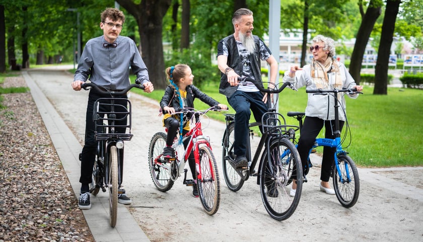 Na zdjęciu grupa rowerzystów w parku, dwóch mężczyzn, dziecko i starsza kobieta 