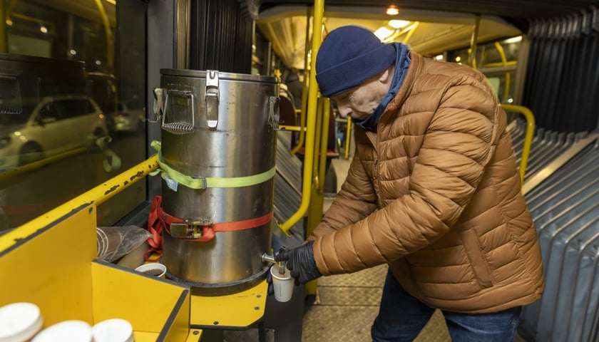 Na zdjęciu mężczyzna w kurtce i czapce, nalewający herbatę z dystrybutora do plastikowego kubka. Wnętrze autobusu "Streetbus", zima 2022. Zdjęcie ilustracyjne