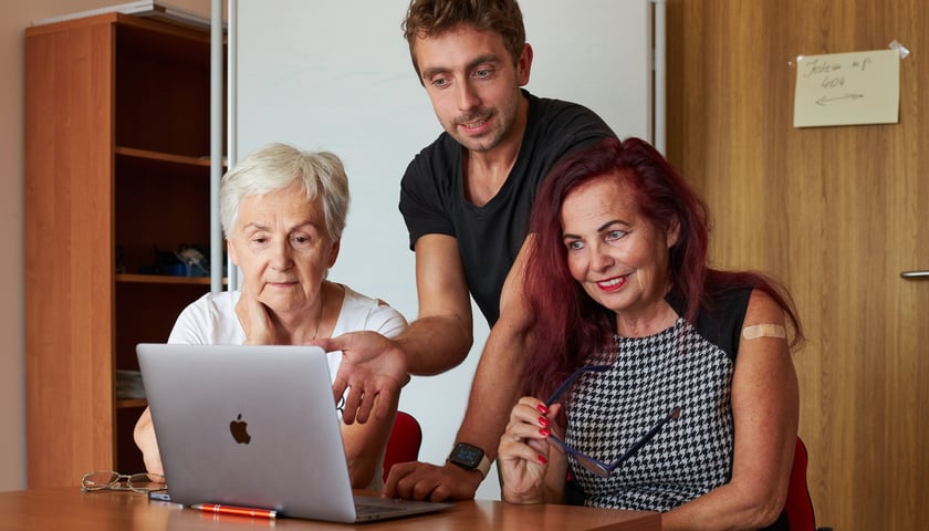 na zdjęciu ilustracyjnym dwie starsze panie siedzą przy stole z komputerem, za plecami stoi młodszy mężczyzna, który coś tłumaczy