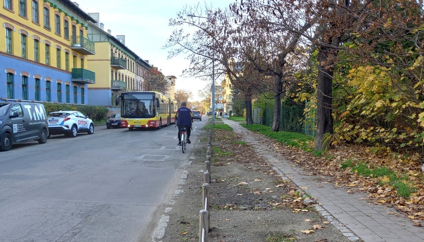 na zdjęciu fragment ulicy, , po lewej stronie stoi blok mieszkalny, pod który parkują samochody, drogą jedzie rowerzysta