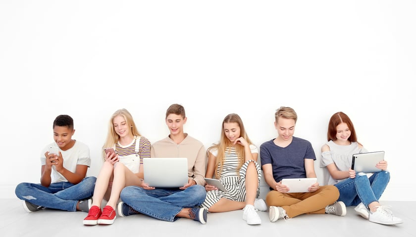 na zdjęciu młodzi ludzie siedzą pod ścianą, trzymają laptopy i czytają z nich