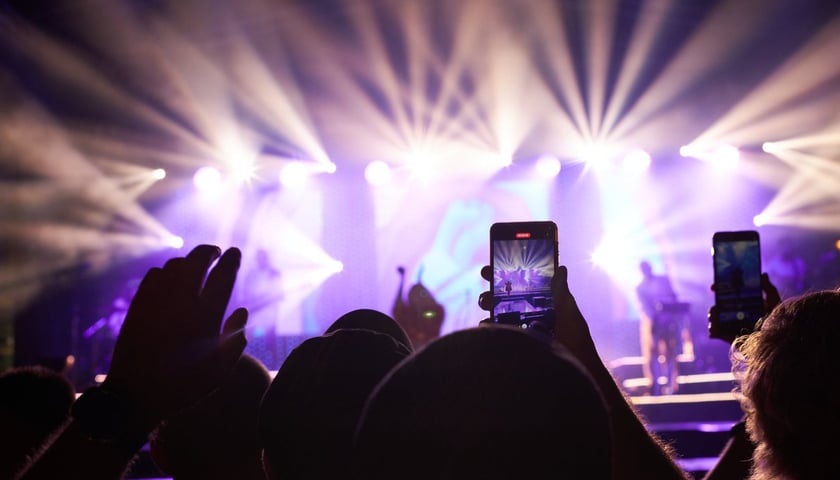 na zdjęciu ilustracyjnym widać podniesione dłonie z telefonami, w tle widać koncert i scenę ze światłami  