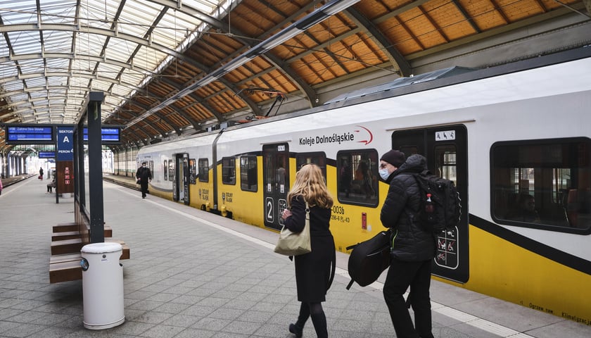 Na zdjęciu widać pociąg Kolei Dolnośląskich, który stoi na dworcu oraz pasażerów na peronie - kobietę i mężczyznę 