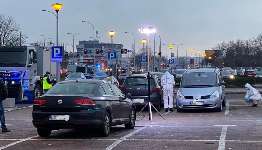 Na zdjęciu widać samochody w miejscu wybuchu na parkingu Centrum Handlowego Marino przy ul. Paprotnej. W tle  - policyjne radiowozy. Obok aut technicy policyjni w białych kombinezonach 