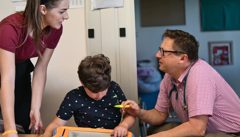 Na zdjęciu dwie dorosłe osoby rozmawiają w obecności dziecka, które rysuje na tablicy magnetycznej. Po prawej prof. Robert Śmigiel, genetyk