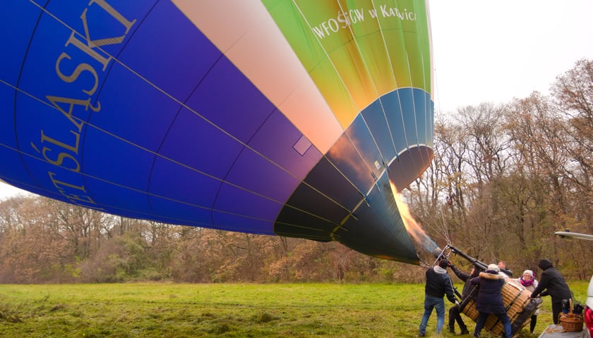 Na zdjęciu widać balon Uniwersytetu Śląskiego w Katowicach wykorzystywany w kampanii pomiarowej PM Cost. Na zdjęciu widać ludzi, którzy przytrzymują balon w momencie napełniania go gorącym powietrzem. 