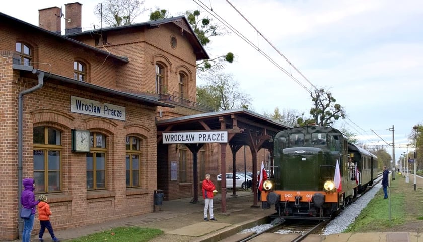 Na zdjęciu widać pociąg retro, który stoi na stacji Wrocław Pracze. Po lewej stronie zdjęcia widać budynki stacji kolejowej z napisem Wrocław Pracze, po prawej - na peronie - zabytkowy  pociąg