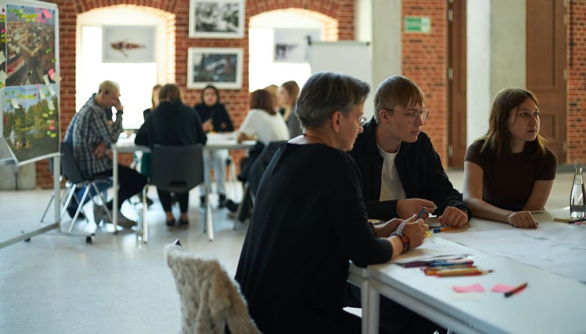 Na zdjęciu dwa stoliki, przy których skupieni są ludzie omawiający rozłożone przed nimi dokumenty. Zdjęcie ilustracyjne, wykonane podczas warsztatów "codzienność w polskiej szkole".