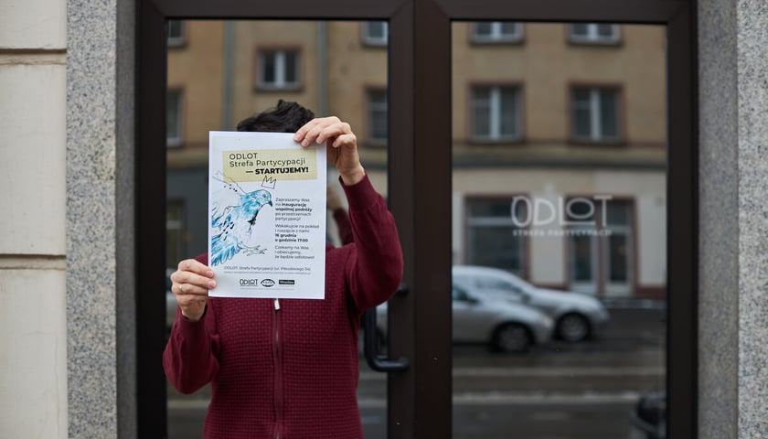 Na zdjęciu wejście do lokalu "ODLOT. Strefa Partycypacji". Człowiek w bordowym swetrze trzyma w rękach plakat informujący o otwarciu ODLOT-u 16 grudnia 2022.