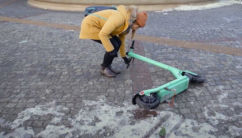 Na kadrze z filmu: kobieta podnosi hulajnogę elektryczną leżącą na placu Orląt Lwowskich