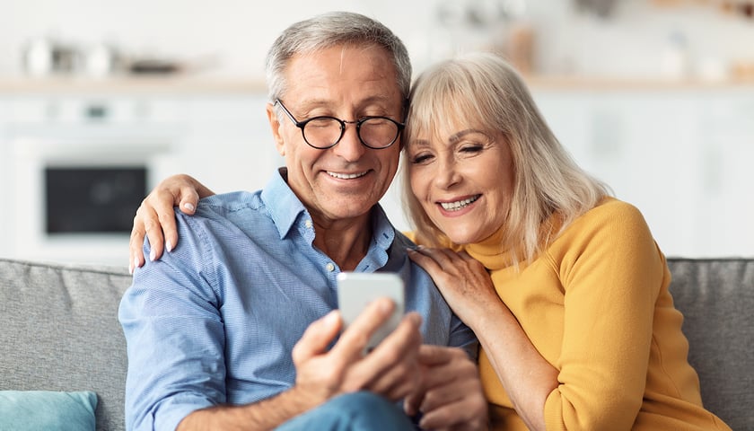 Na zdjęciu widać starszą kobietę i mężczyznę patrzących na telefon trzymany w dłoni
