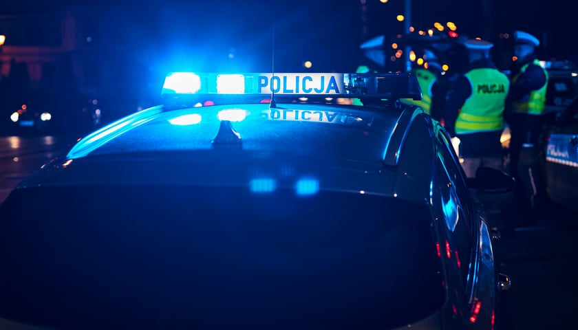 Na zdjęciu policyjny radiowóz nocą, w tle funkcjonariusze policji / zdjęcie ilustracyjne