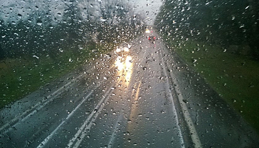 Na zdjęciu zasnuta kroplami deszczu szyba, przez którą widać drogę i przejeżdżający nią samochód. Zdjęcie ilustracyjne