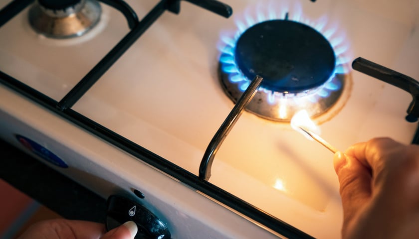 Na zdjęciu widać zapalony palnik w kuchence gazowej i dłoń z zapaloną zapałką 