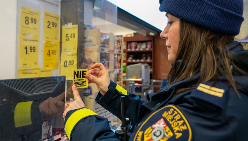 Na zdjęciu strażniczka miejska nakleja na szybie sklepu naklejkę z napisem "Nie sprzedawaj alkoholu mojemu dziecku"