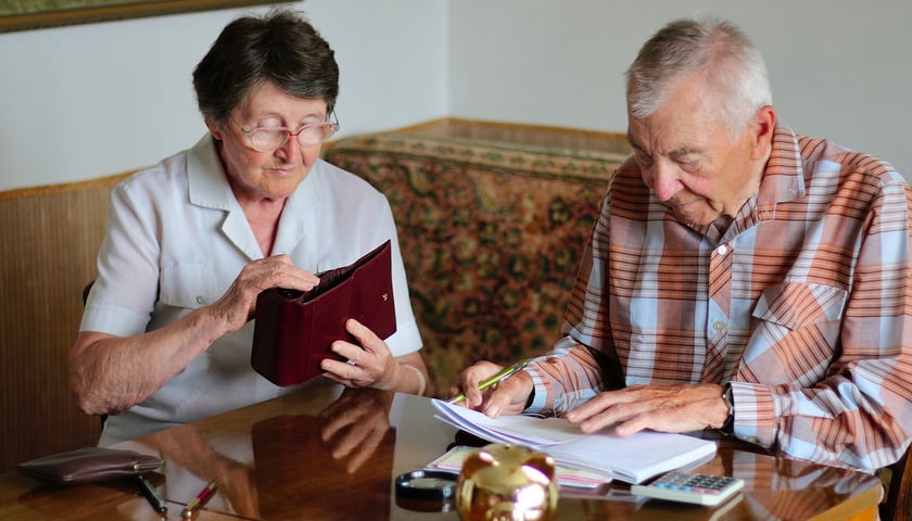 Na zdjęciu widać seniorów: kobietę i mężczyznę siedzących przy stole, na którym leżą pieniądze i dokumenty 
