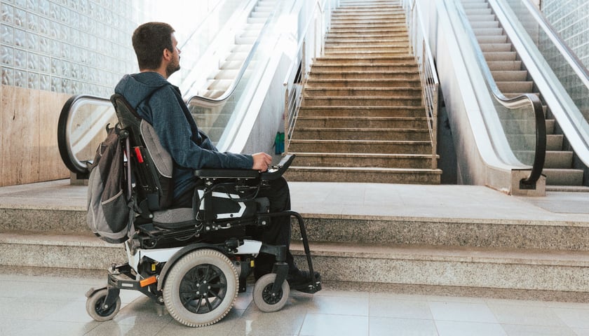 Na zdjęciu mężczyzna na wózku inwalidzkim spoglądający na wysokie schody, będące dla niego barierą architektoniczną. Zdjęcie ilustracyjne.