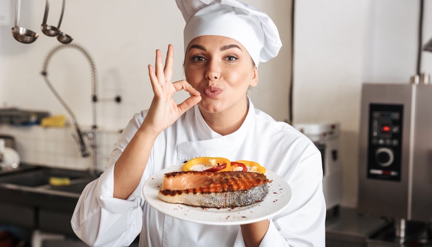 Na zdjęciu uśmiechnięta szefowa kuchni pokazująca biały talerz z pieczoną rybą