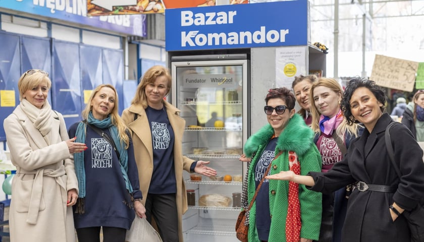 Otwarcie jadłodzielni na Bazarze Komandor. Na zdjęciu widać siedem uśmiechniętych kobiet, stojących po bokach nowej lodówki do dzielenia się produktami spożywczymi.