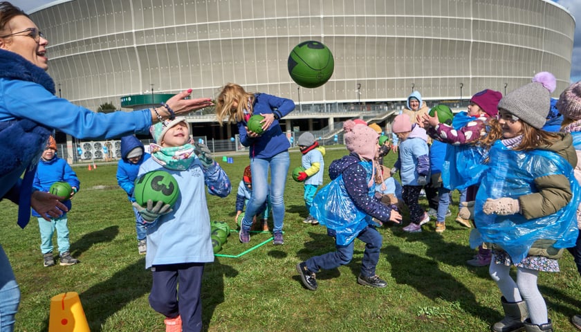 Dzieci i opiekunki bawią się piłką i skaczą, w tle stadion