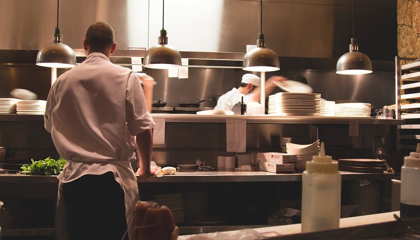 Restauracyjna kuchnia. W pomieszczeniu jest dwóch kucharzy. Jeden stoi tyłem przy blacie roboczym, drugi, w głębi kuchni, sięga ręką po talerz (zdjęcie ilustracyjne)