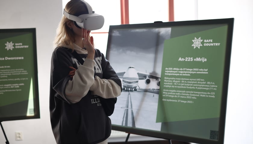 Kobieta w okularach do wirtualnej rzeczywistości stoi między dwiema dużymi tablicami informacyjnymi  
