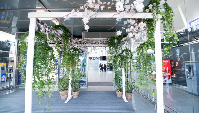 Wejście przystrojone jasnymi kwiatami, w tle: osoby w terminalu lotniska