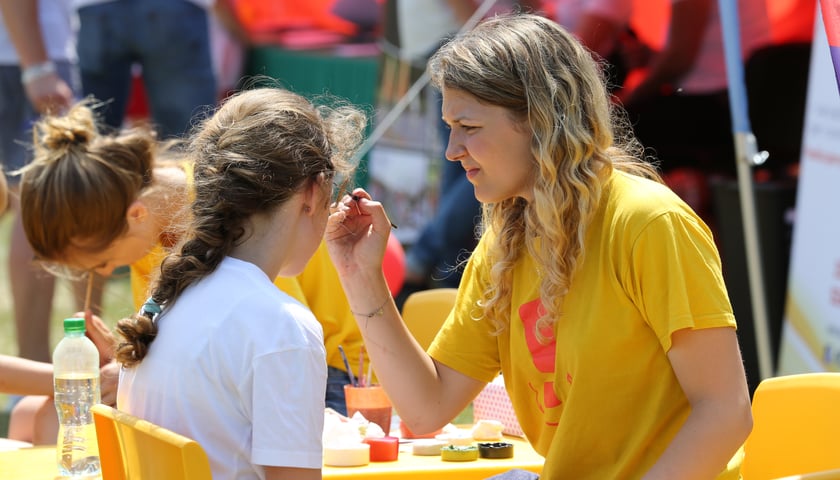 Kobieta w żółtej koszulce maluje twarz dziewczynce w białej koszulce