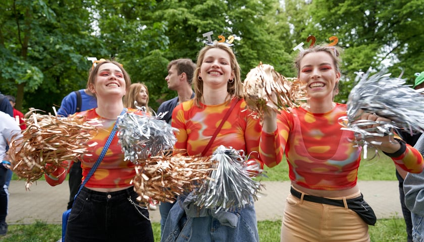 Trzy uśmiechnięte cheerleaderki ubrane w pomarańczowe bluzki, w dłoniach trzymają złote i srebrne pomponami z folii, a na głowie mają opaski z literami T 22
