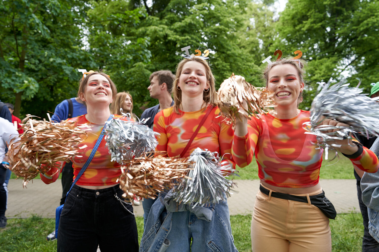 Trzy uśmiechnięte cheerleaderki ubrane w pomarańczowe bluzki, w dłoniach trzymają złote i srebrne pomponami z folii, a na głowie mają opaski z literami T 22