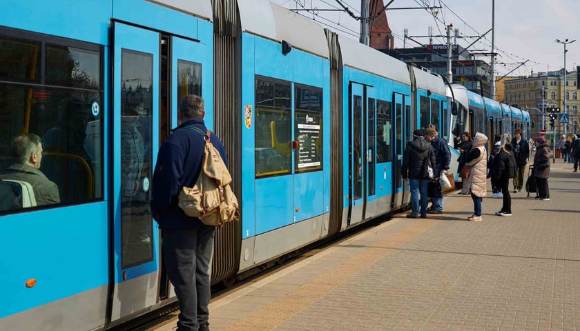 Niebieski tramwaj na przystanku oraz czekający na wejście do niego pasażerowie
