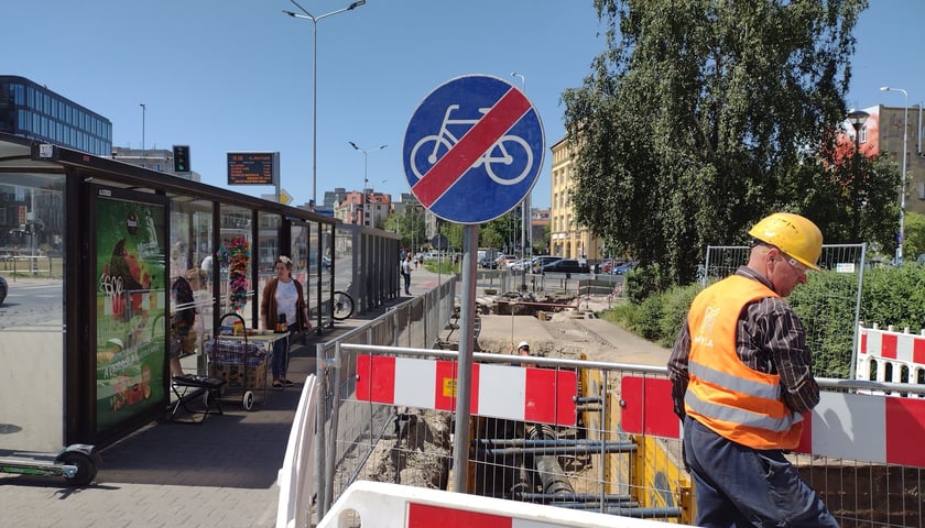 Rów w drodze rowerowej, przekreślony znak z symbolem roweru, mężczyzna w kamizelce i kasku, a także tylna strona wiaty przystankowej