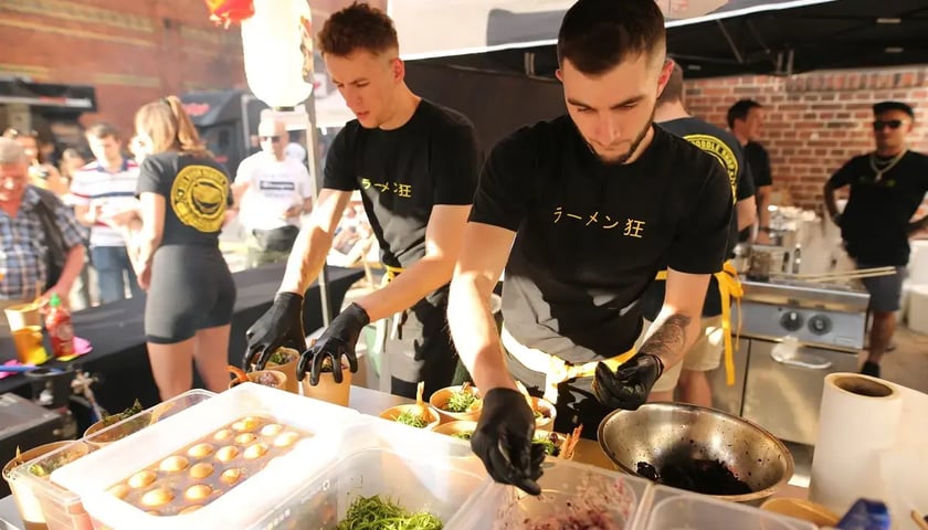 Dwaj mężczyźni w czarnych podkoszulkach i lateksowych rękawiczkach nakładający jedzenie w namiocie podczas imprezy Gastro Miasto