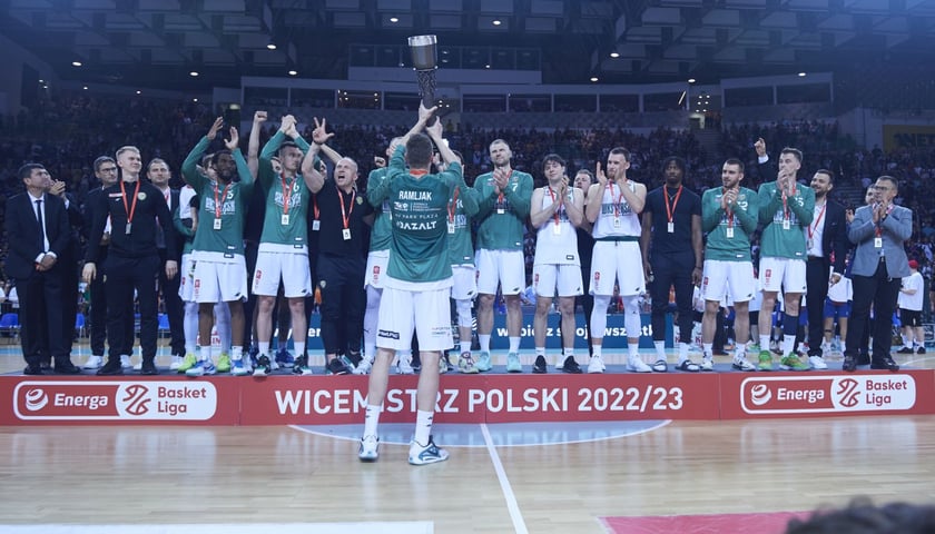 Ivan Ramljak unosi puchar za drugie miejsce. Pozostali koszykarze i inni członkowie drużyny stoją na podium