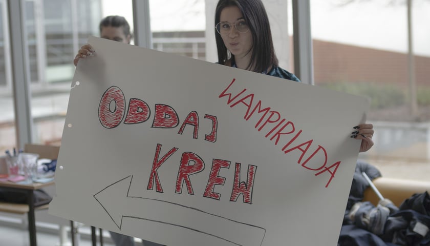 Młoda kobieta w okularach trzyma dużą arkusz papieru z odręcznie wykonanym napisem: Wampiriada. Oddaj krew. Narysowana jest też strzałka. Plakat z Wapiriady, która miała miejsce w lutym 2023 r.  