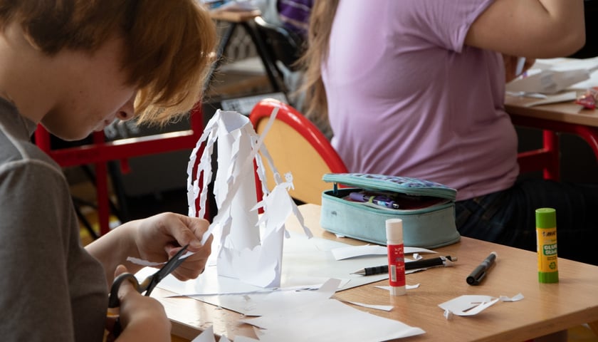 Młoda osoba tnie nożyczkami papier. Przed nią na stole widać nieporządek: dużo papieru, konstrukcję przestrzenną z papieru, tubkę kleju, piórnik. W tle tyłem siedzi osoba  