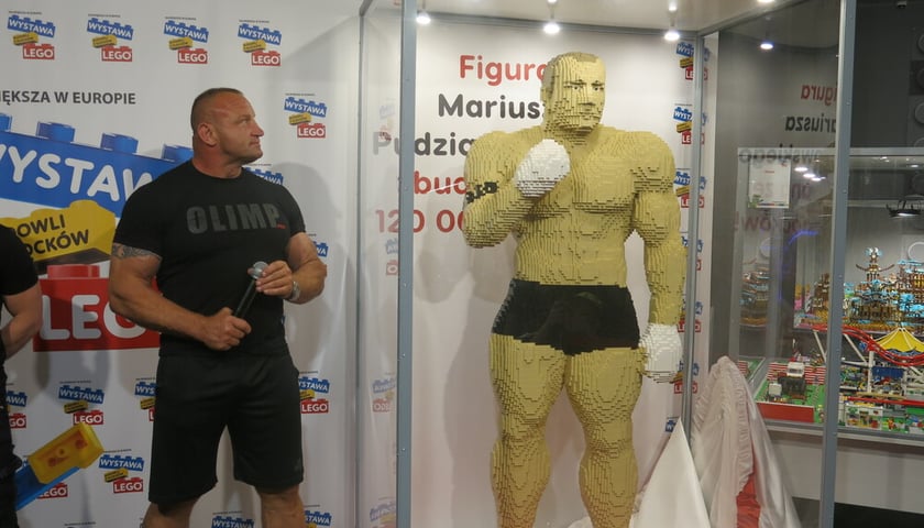 Po lewej umięśniony mężczyzna ubrany w czarną koszulkę i spodenki  - Mariusz Pudzianowski - patrzy na figurę z żółtych klocków lego, która stoi po prawej stronie. Figura takiej samej wielkości jak Pudzianowski umieszczona jest w szklanej gablocie. 