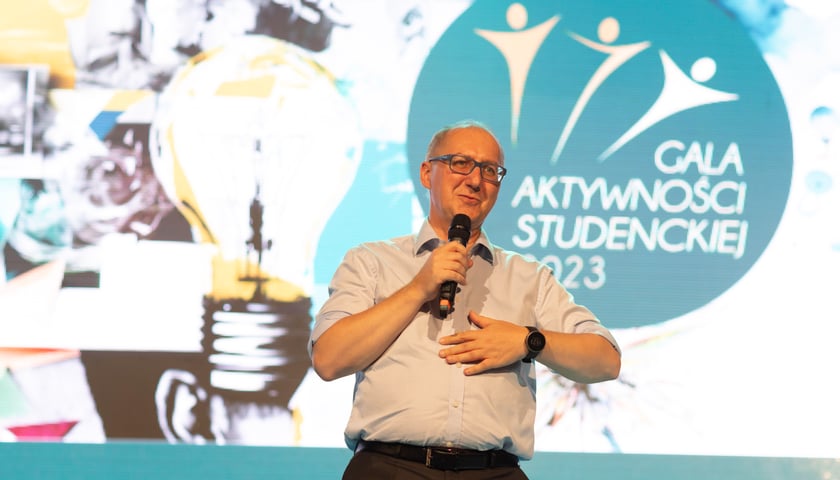 Mężczyzna w jasnej koszuli z mikrofonem w dłoni: prof. Arkadiusz Wójs, rektor Politechniki Wrocławskiej; za nim grafika z żarówką i napisem: Gala Aktywności Studenckiej 2023 